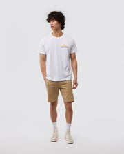 A person stands against a white background, wearing the MENS CALIFORNIA TEE - B6U552W1PC by Psycho Bunny, made from breathable Pima cotton jersey. They pair it with beige shorts, white socks, and white sneakers. The t-shirt features a small graphic on the left chest area. The person has curly hair and a relaxed posture.