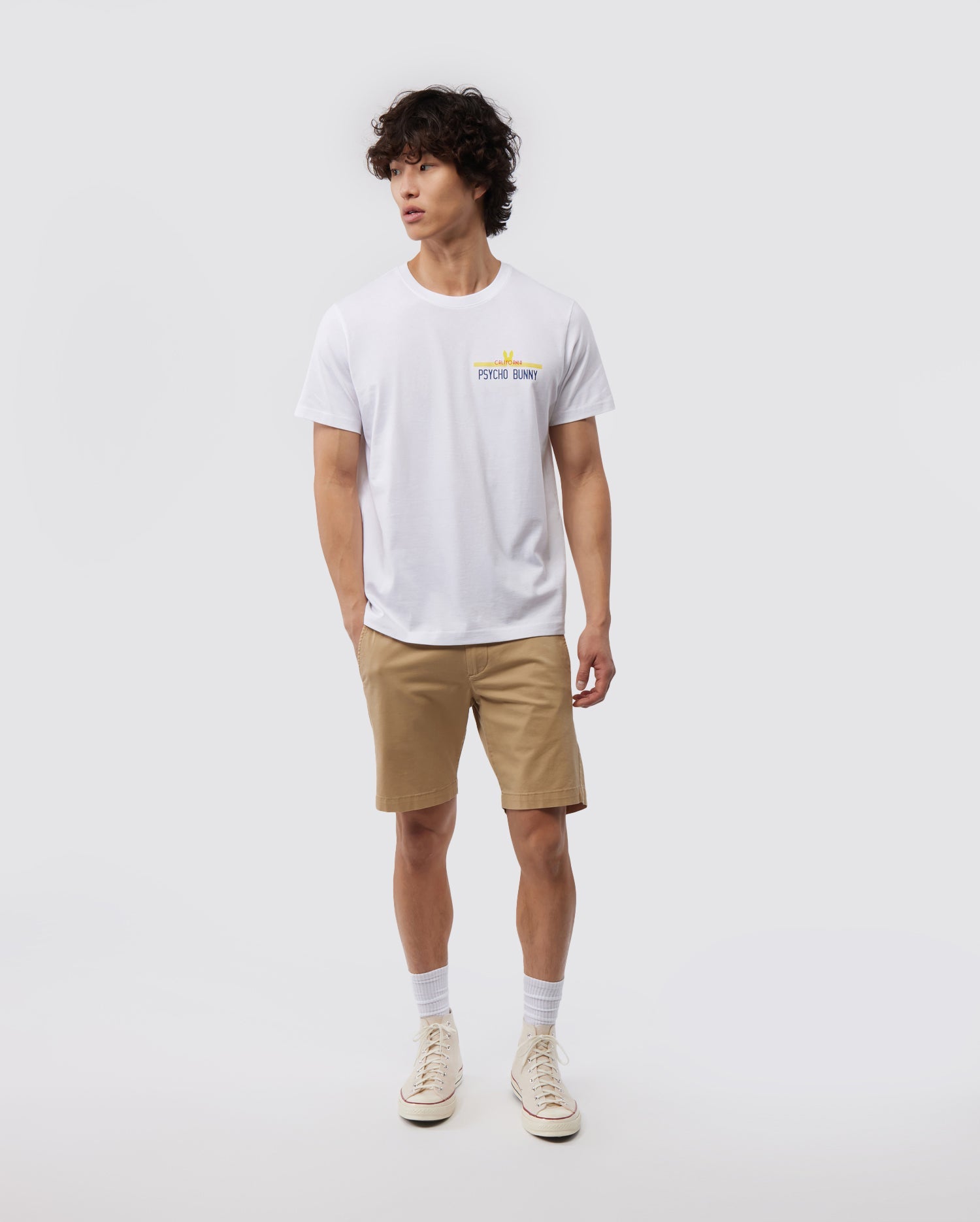 A person stands against a white background, wearing the MENS CALIFORNIA TEE - B6U552W1PC by Psycho Bunny, made from breathable Pima cotton jersey. They pair it with beige shorts, white socks, and white sneakers. The t-shirt features a small graphic on the left chest area. The person has curly hair and a relaxed posture.