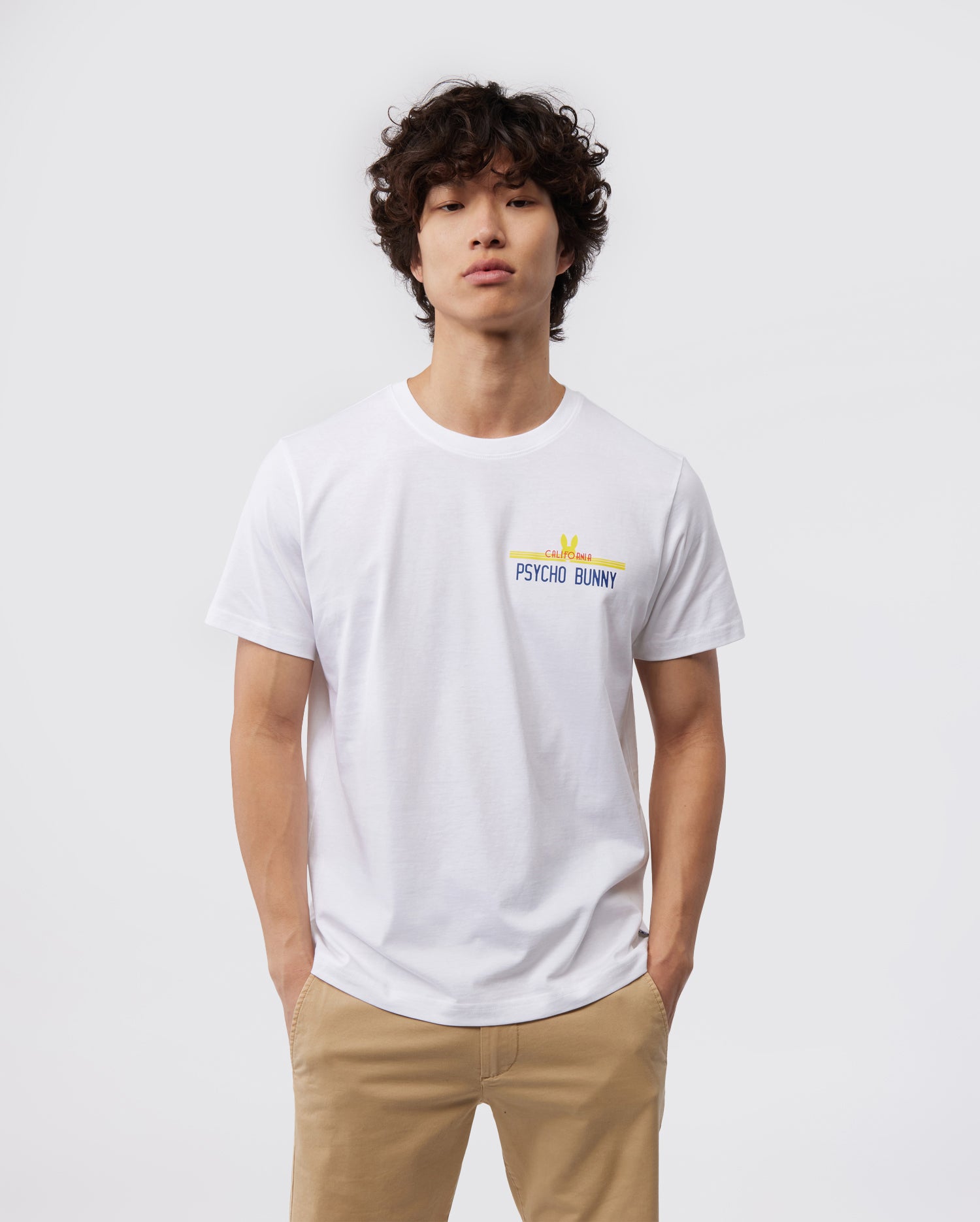 A person with curly hair is wearing a clean, white MENS CALIFORNIA TEE - B6U552W1PC from Psycho Bunny, made of breathable Pima cotton jersey. The tee features the "Psycho Bunny" logo and a small yellow rabbit above the text. They are standing against a plain, light gray background with both hands in the pockets of their tan pants.