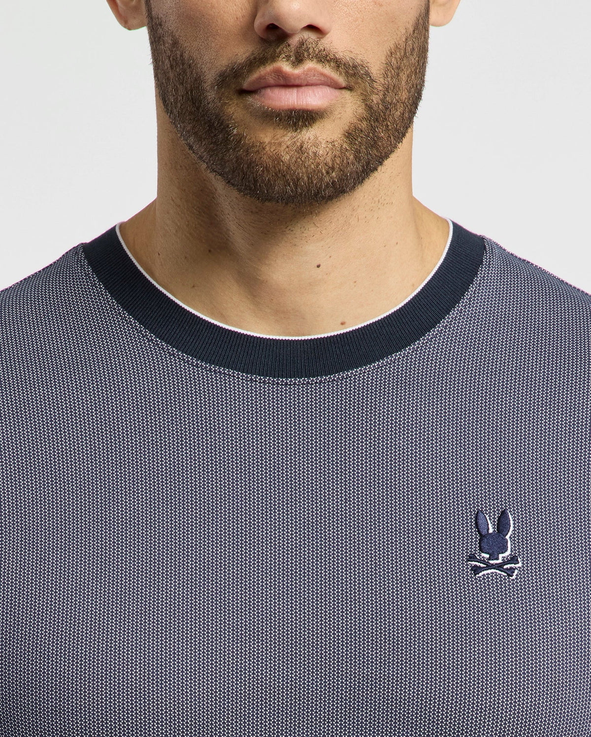 A man wears the Psycho Bunny Men's Mackay Jacquard Fashion Tee, featuring a premium dark gray textured design with a black collar. Made from Jacquard-knit Pima cotton, it showcases an embroidered chest Bunny logo with crossed bones on the left against a white background.