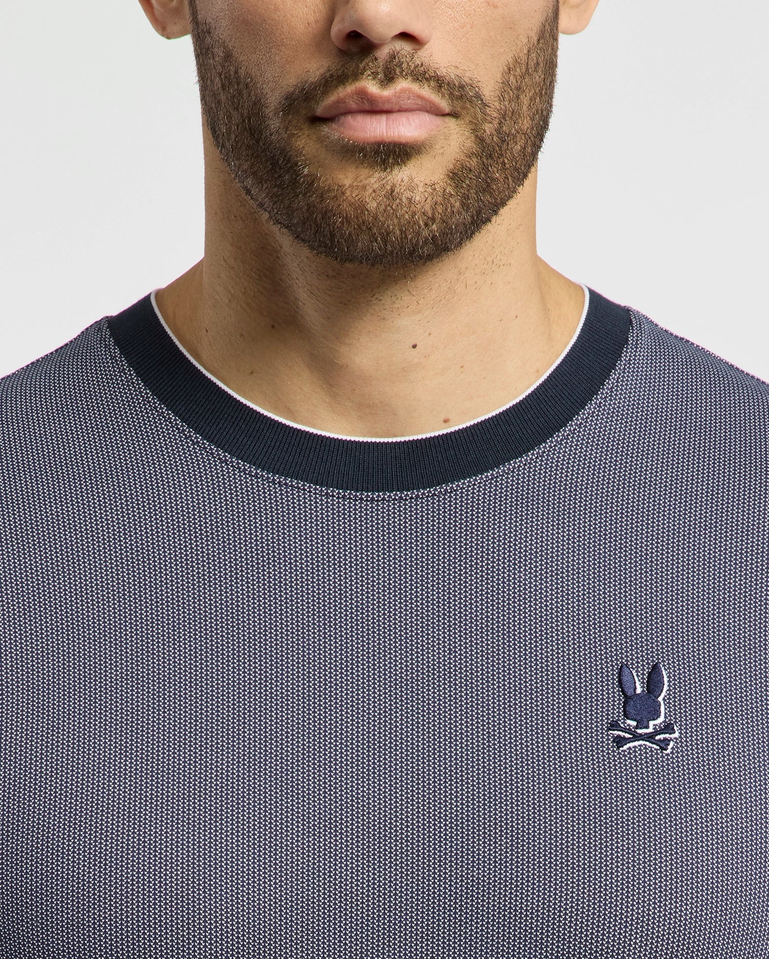 A man wears the Psycho Bunny Men's Mackay Jacquard Fashion Tee, featuring a premium dark gray textured design with a black collar. Made from Jacquard-knit Pima cotton, it showcases an embroidered chest Bunny logo with crossed bones on the left against a white background.