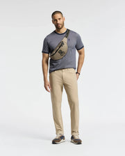 A man models the Psycho Bunny MENS MACKAY JACQUARD FASHION TEE (B6U576F200) in blue with beige pants and a crossbody bag featuring an embroidered Bunny. He stands against a white background, wearing sneakers, looking at the camera.