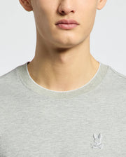 A person is dressed in the MENS MACKAY JACQUARD FASHION TEE - B6U576F200 from Psycho Bunny. The tee is light gray, Pima cotton, and features an embroidered white bunny logo on the chest against a plain white background.
