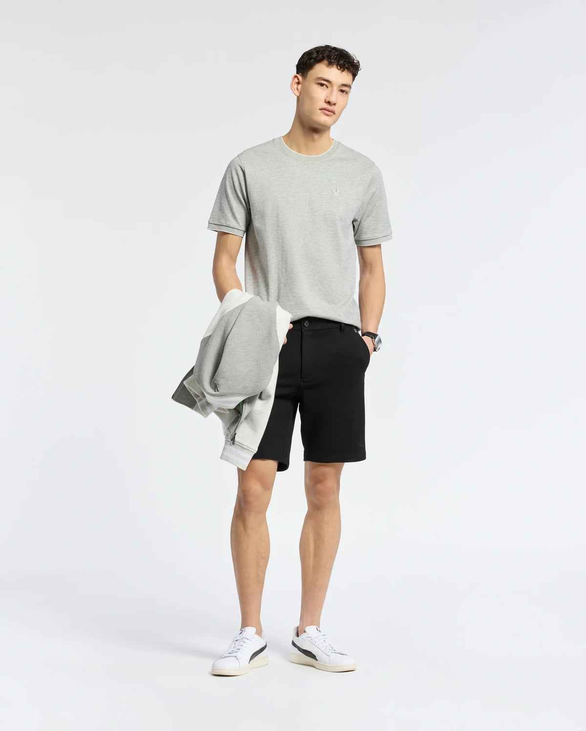 A person stands against a plain background wearing a Psycho Bunny light gray Pima cotton tee paired with black shorts, holding a matching embroidered chest Bunny sweatshirt. They have short hair and white sneakers, gazing at the camera with a neutral expression.
