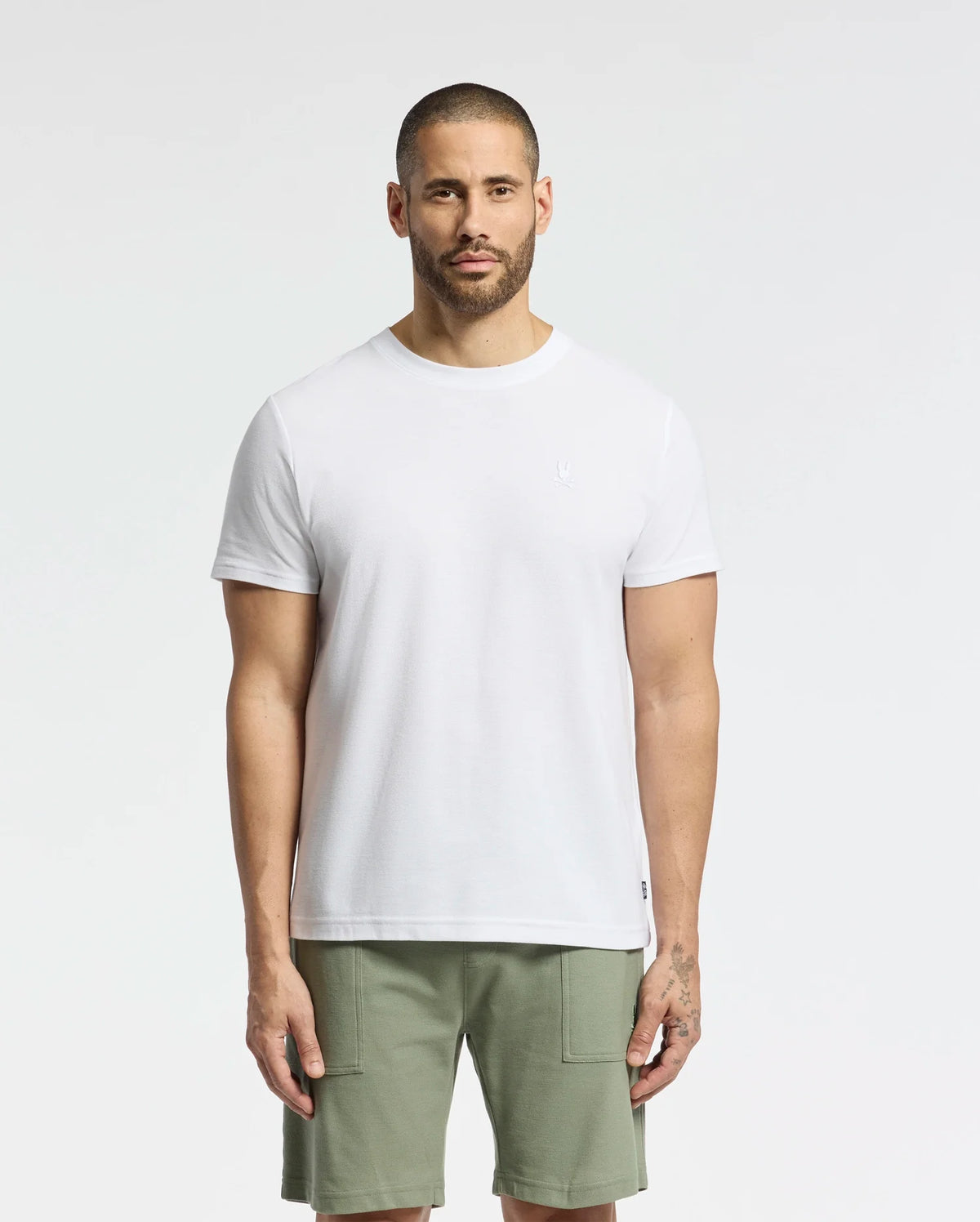 A short-haired, bearded individual stands against a white backdrop wearing a Psycho Bunny MENS TAYLOR PIQUE TEE in regular fit, crafted from lightweight Pima cotton, complemented by olive green shorts. Their relaxed stance is highlighted by the tee's embroidered chest bunny.
