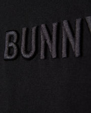 Close-up of the word "Psycho Bunny" embroidered logo in black thread on a black fabric background, with a focus on texture and detail.