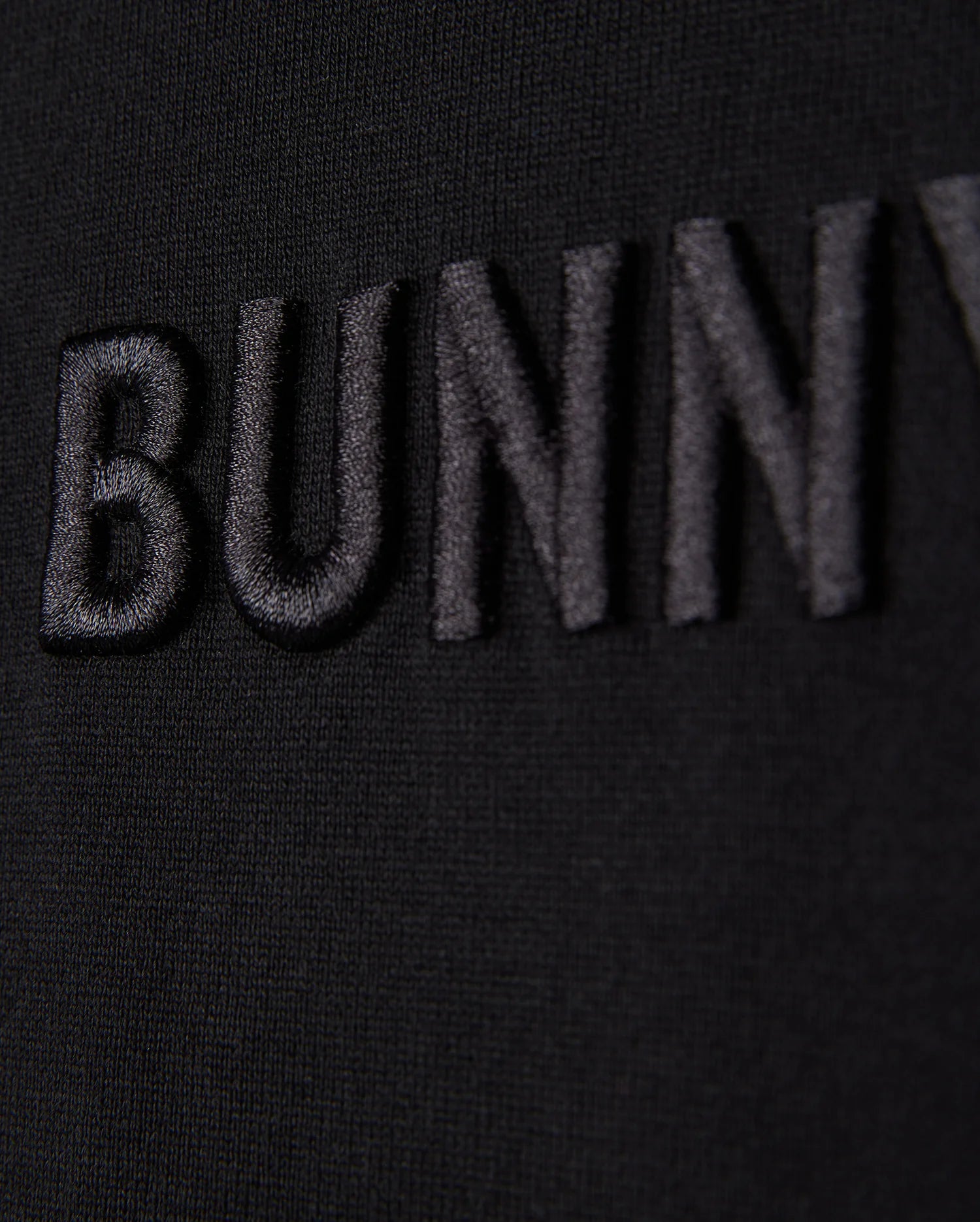 Close-up of the word "Psycho Bunny" embroidered logo in black thread on a black fabric background, with a focus on texture and detail.