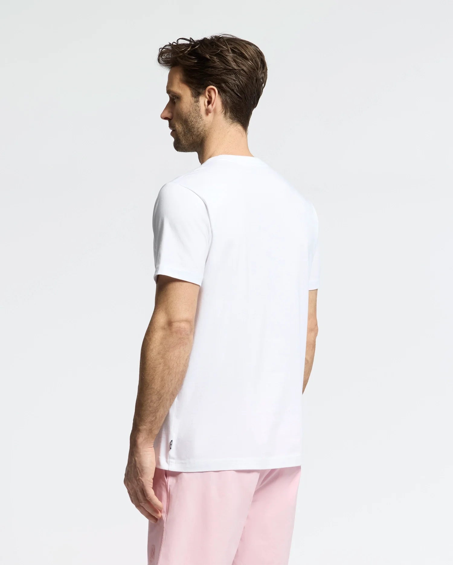 A brown-haired man wears a white Psycho Bunny MENS GOLDIE GRAPHIC TEE - B6U606F200 with a subtle bunny graphic, paired with pink pants. He stands with his back turned, looking left against a plain, light background.