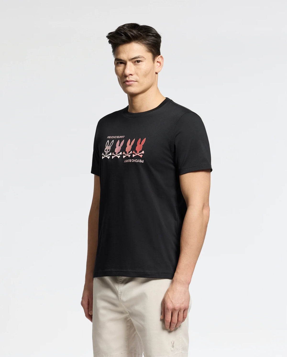 A person wearing Psycho Bunny's MENS OCEANIA EMBROIDERED GRAPHIC TEE - B6U607F200 stands against a plain white background. The black Pima cotton tee features retro embroidery with pink and red bunny outlines, while the person wears beige pants and looks toward the camera.