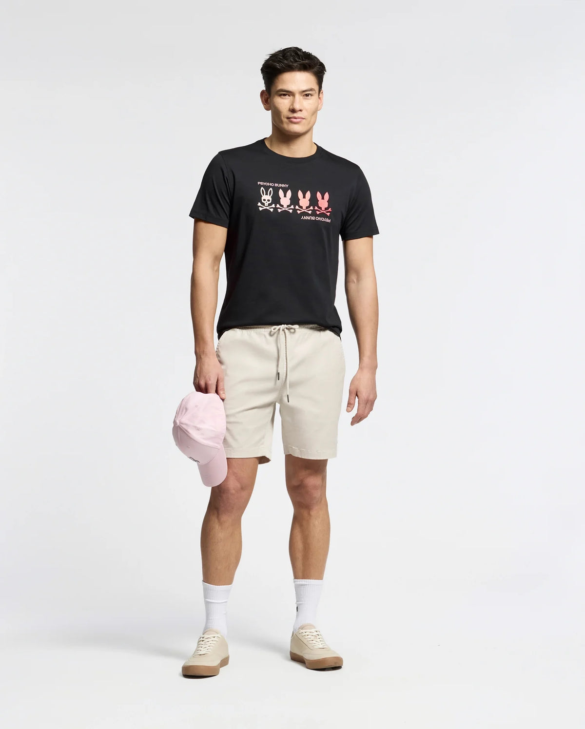 A man stands against a plain background wearing a Psycho Bunny black Pima Cotton Tee featuring an embroidered bunny, beige shorts, white socks, and light brown sneakers. He holds a light pink baseball cap at his side.