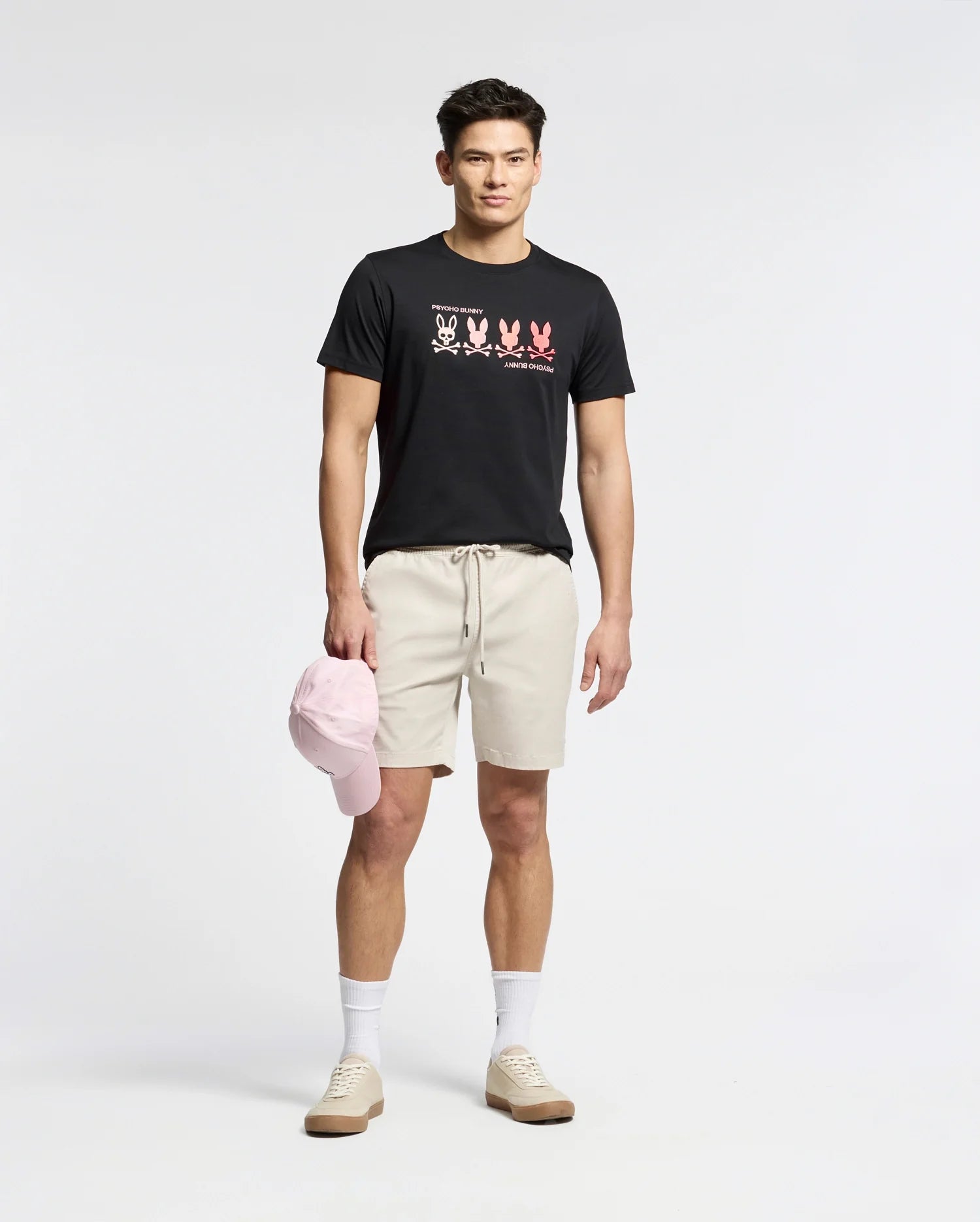 A man stands against a plain background wearing a Psycho Bunny black Pima Cotton Tee featuring an embroidered bunny, beige shorts, white socks, and light brown sneakers. He holds a light pink baseball cap at his side.