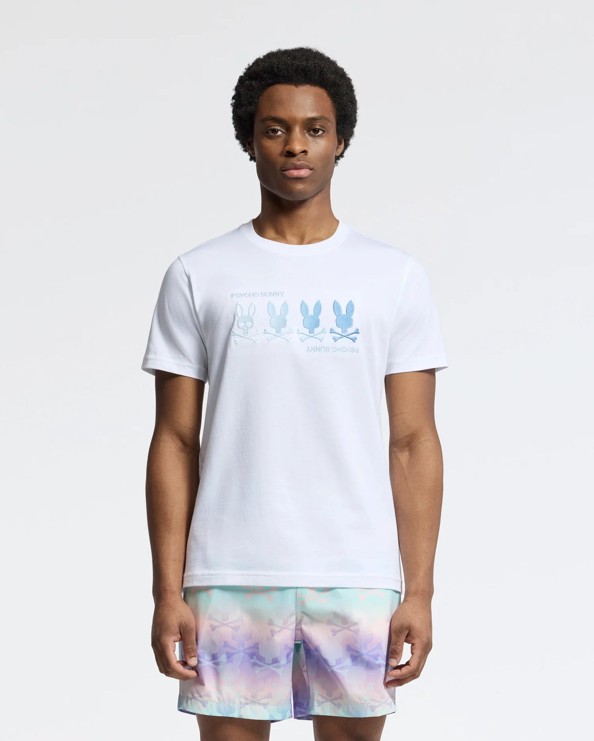 A person stands wearing a Psycho Bunny Men's Oceania Embroidered Graphic Tee (B6U607F200), featuring blue bunny graphics with text, and colorful pastel shorts against a plain white background.