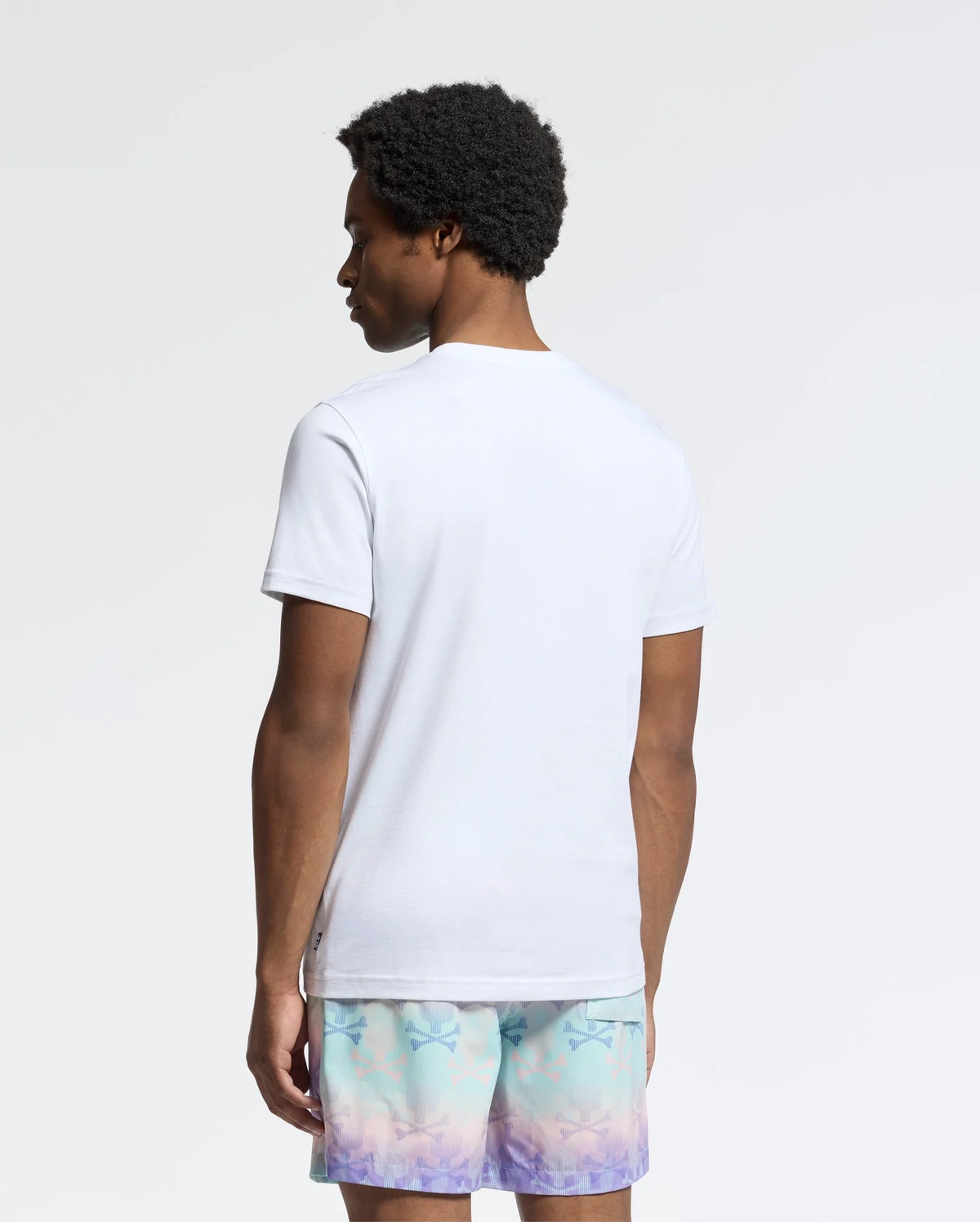 A person with curly hair stands facing away, wearing a Psycho Bunny Men's Oceania Embroidered Graphic Tee and pastel shorts. The plain, light-colored background enhances the retro vibes of the effortlessly cool scene.