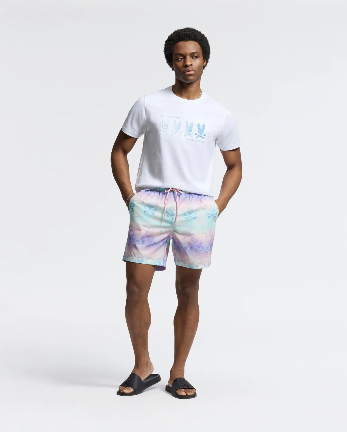 A man stands relaxed with hands in pockets, wearing Psycho Bunny's white MENS OCEANIA EMBROIDERED GRAPHIC TEE - B6U607F200 with a bunny design, pastel tie-dye shorts, and black slides, all exuding retro vibes against a plain light gray backdrop.