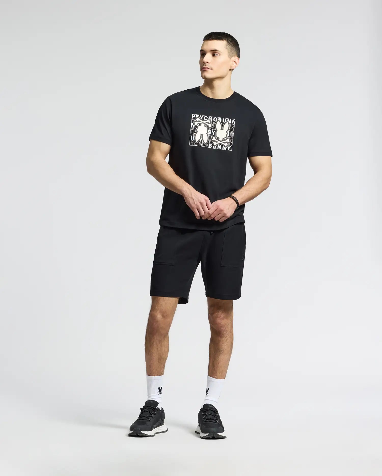 A man wearing a Psycho Bunny MENS GOLD COAST GRAPHIC TEE in black, paired with black shorts, stands against a plain backdrop. He completes the look with black sneakers and white socks featuring a logo, showcasing premium craftsmanship. His clasped hands embody timeless style effortlessly.