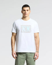 A person is wearing a Psycho Bunny MENS GOLD COAST GRAPHIC TEE - B6U631F200, a premium Pima cotton white t-shirt with abstract figures in a square, paired with green pants. The classic fit enhances the simple light background elegantly.