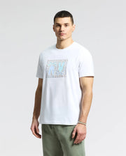 Against a plain backdrop, a person wears the Psycho Bunny MENS GOLD COAST GRAPHIC TEE - B6U631F200, made from premium Pima cotton and featuring blue abstract figures. They pair it with classic fit green pants and a bracelet, maintaining a neutral expression.