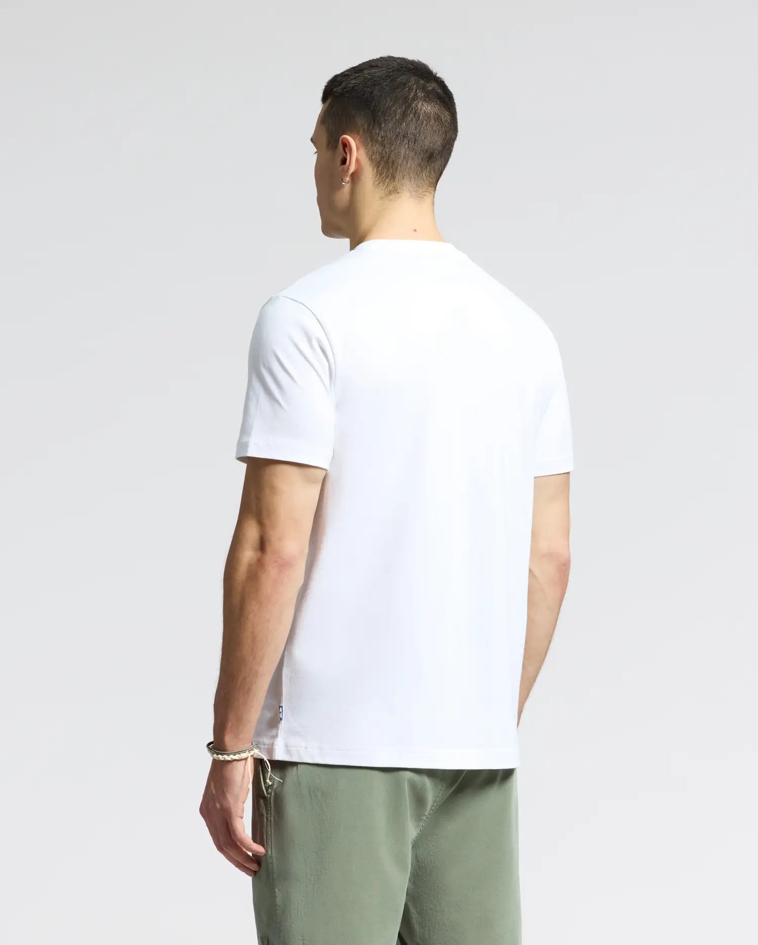 A man with short hair has his back to the camera, wearing a plain white MENS GOLD COAST GRAPHIC TEE by Psycho Bunny and green shorts. The light gray background highlights the classic fit of his attire.