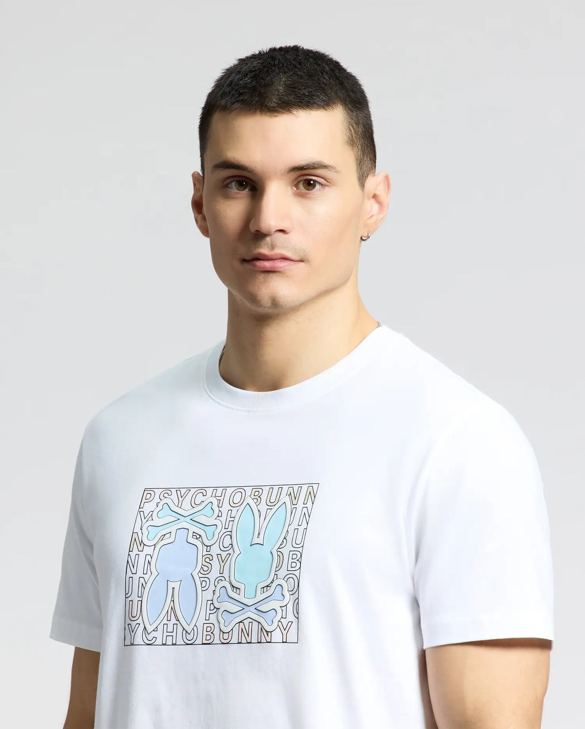 A man in a classic fit, white Psycho Bunny Gold Coast Graphic Tee made of premium Pima cotton, featuring a blue bunny graphic with crossbones and "PSYCHO BUNNY" text, stands with short dark hair against a neutral backdrop, radiating understated style and quality craftsmanship.
