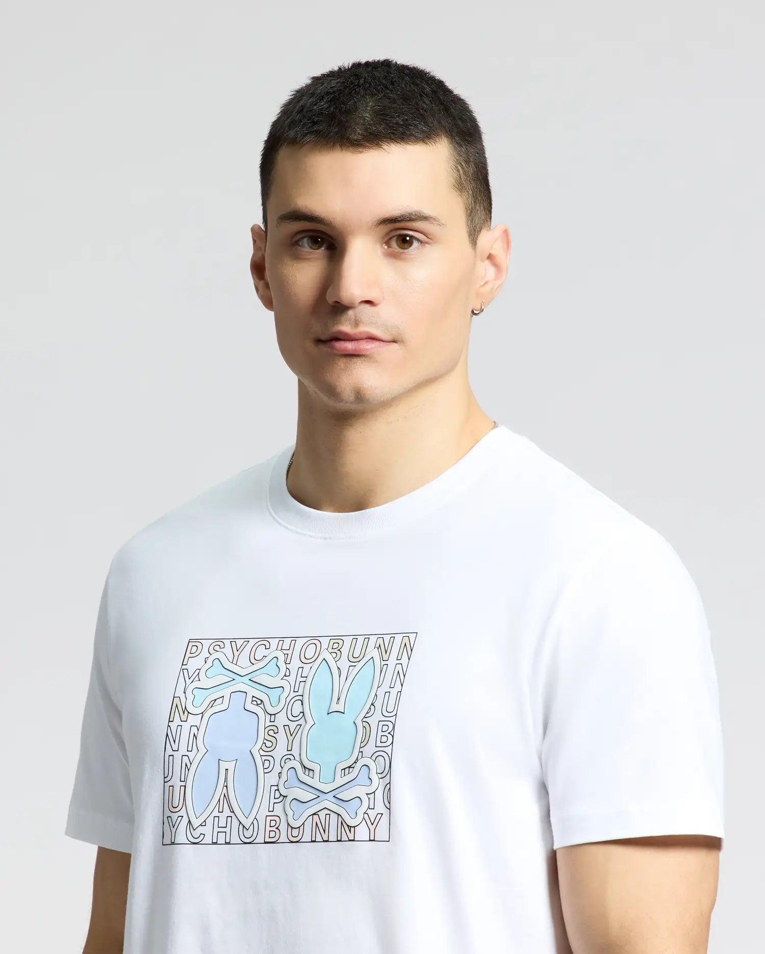 A man in a classic fit, white Psycho Bunny Gold Coast Graphic Tee made of premium Pima cotton, featuring a blue bunny graphic with crossbones and "PSYCHO BUNNY" text, stands with short dark hair against a neutral backdrop, radiating understated style and quality craftsmanship.