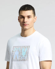 A man in a classic fit, white Psycho Bunny Gold Coast Graphic Tee made of premium Pima cotton, featuring a blue bunny graphic with crossbones and "PSYCHO BUNNY" text, stands with short dark hair against a neutral backdrop, radiating understated style and quality craftsmanship.