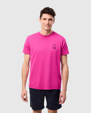 A man stands against a plain background wearing the MENS PEARLAND BACK GRAPHIC TEE - B6U632C200 from Psycho Bunny in vibrant pink, featuring a small embroidered bunny logo on the left side of his chest. Made from luxurious Pima cotton, the shirt perfectly complements his short dark hair and cheerful smile. He is also dressed in dark shorts.