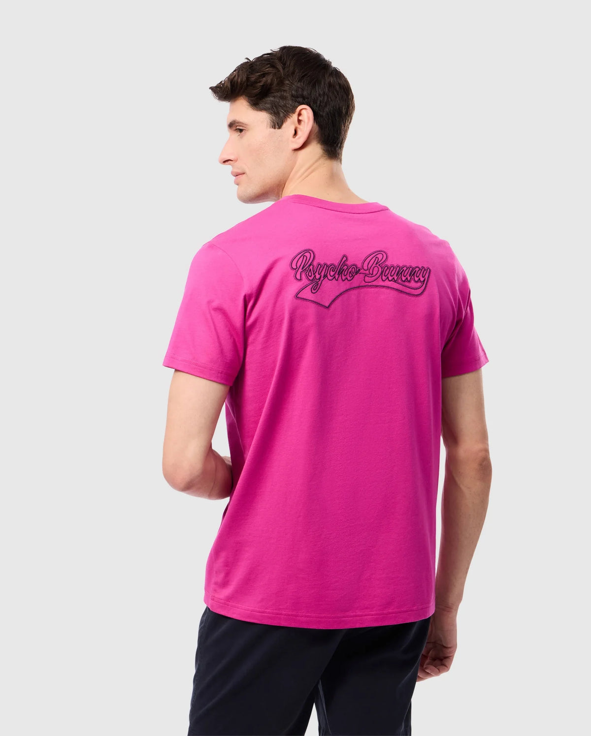 A man with short brown hair, wearing a bright pink Psycho Bunny MENS PEARLAND BACK GRAPHIC TEE – B6U632C200 made from soft Pima cotton and black shorts, stands facing backward with his head slightly turned to the side. The back of the tee features embroidered cursive text. The background is a plain light gray.