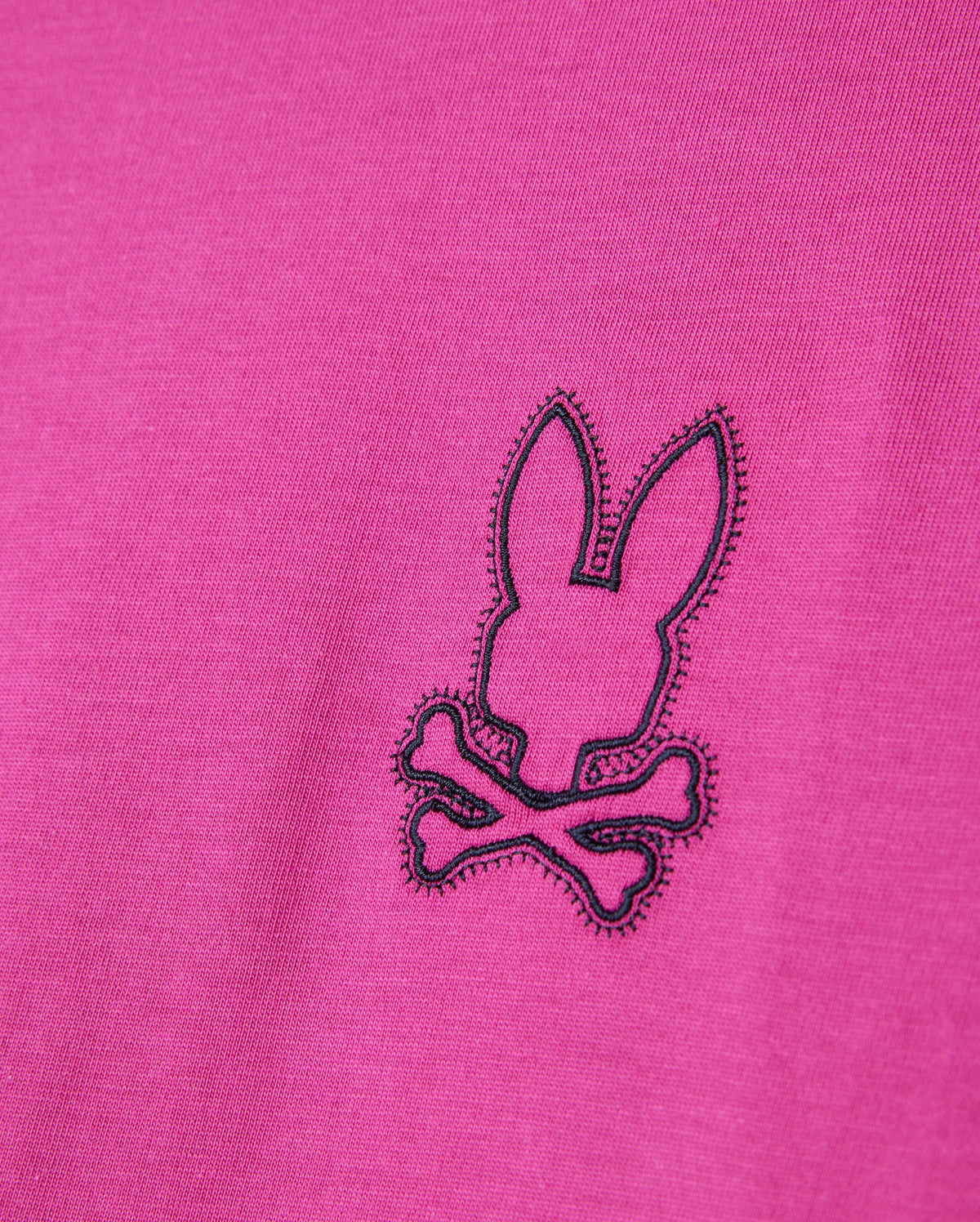Close-up of the Psycho Bunny MEN'S PEARLAND BACK GRAPHIC TEE - B6U632C200 in pink, showcasing an embroidered black outline of a bunny head above two crossed bones, creating a playful pirate-inspired logo. The stitching is intricate, offering a textured contrast to the Pima cotton fabric.