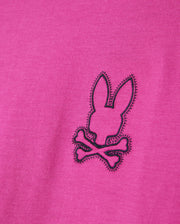 Close-up of the Psycho Bunny MEN'S PEARLAND BACK GRAPHIC TEE - B6U632C200 in pink, showcasing an embroidered black outline of a bunny head above two crossed bones, creating a playful pirate-inspired logo. The stitching is intricate, offering a textured contrast to the Pima cotton fabric.
