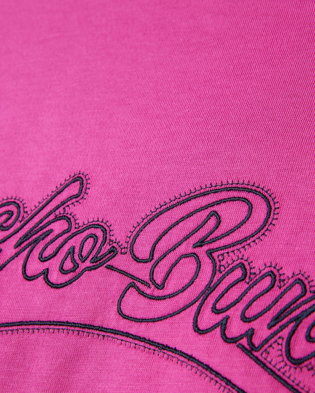A close-up of the Psycho Bunny MENS PEARLAND BACK GRAPHIC TEE - B6U632C200 in soft pink Pima cotton, showcasing intricate black embroidered cursive letters. The bold and heavy embroidery contrasts sharply against the vivid pink background, with detailed stitching and fabric texture clearly visible.