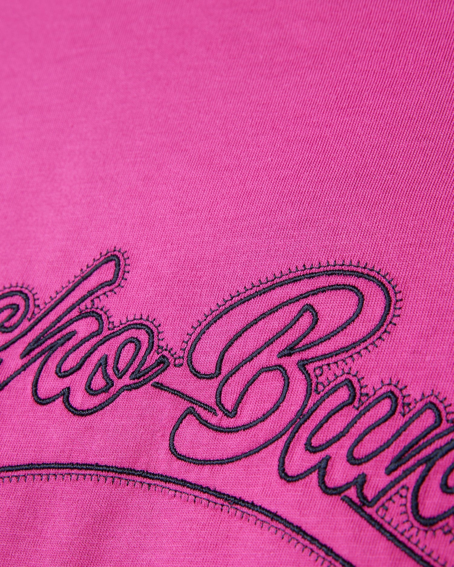 A close-up of the Psycho Bunny MENS PEARLAND BACK GRAPHIC TEE - B6U632C200 in soft pink Pima cotton, showcasing intricate black embroidered cursive letters. The bold and heavy embroidery contrasts sharply against the vivid pink background, with detailed stitching and fabric texture clearly visible.