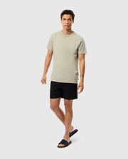 A person wearing a light beige MENS PEARLAND BACK GRAPHIC TEE - B6U632C200 from Psycho Bunny, black shorts, and black slide sandals is standing against a plain white background. The embroidered Pima cotton shirt contributes to the casual and relaxed vibe, with one hand slightly extended and the other by their side.