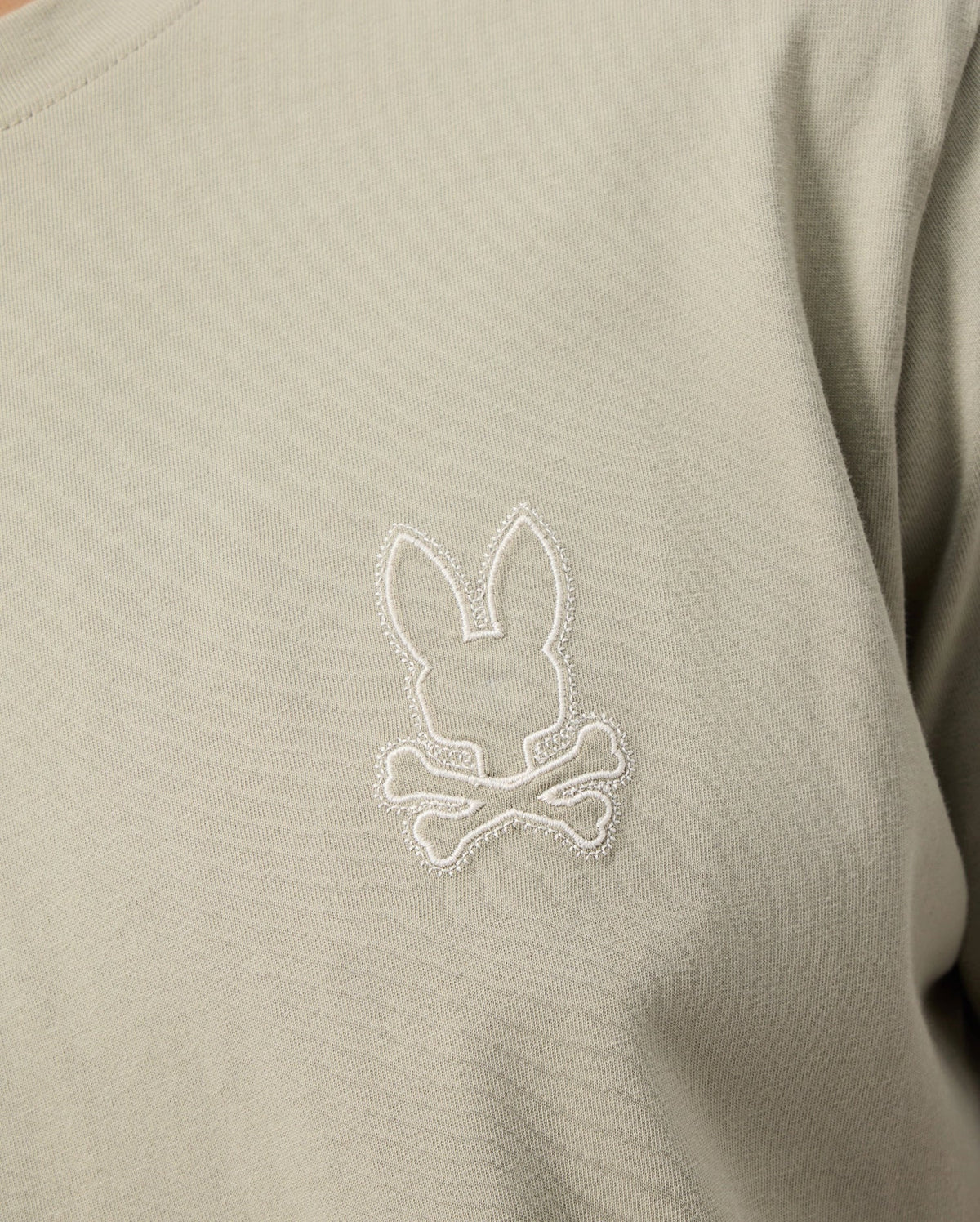 A close-up image of the Psycho Bunny MENS PEARLAND BACK GRAPHIC TEE - B6U632C200 in light grey-green, featuring an embroidered rabbit head above a pair of crossed bones on the chest. The intricate stitching is done in white, creating a chic contrast with the Pima cotton fabric.