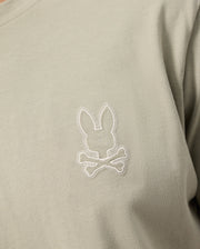 A close-up image of the Psycho Bunny MENS PEARLAND BACK GRAPHIC TEE - B6U632C200 in light grey-green, featuring an embroidered rabbit head above a pair of crossed bones on the chest. The intricate stitching is done in white, creating a chic contrast with the Pima cotton fabric.