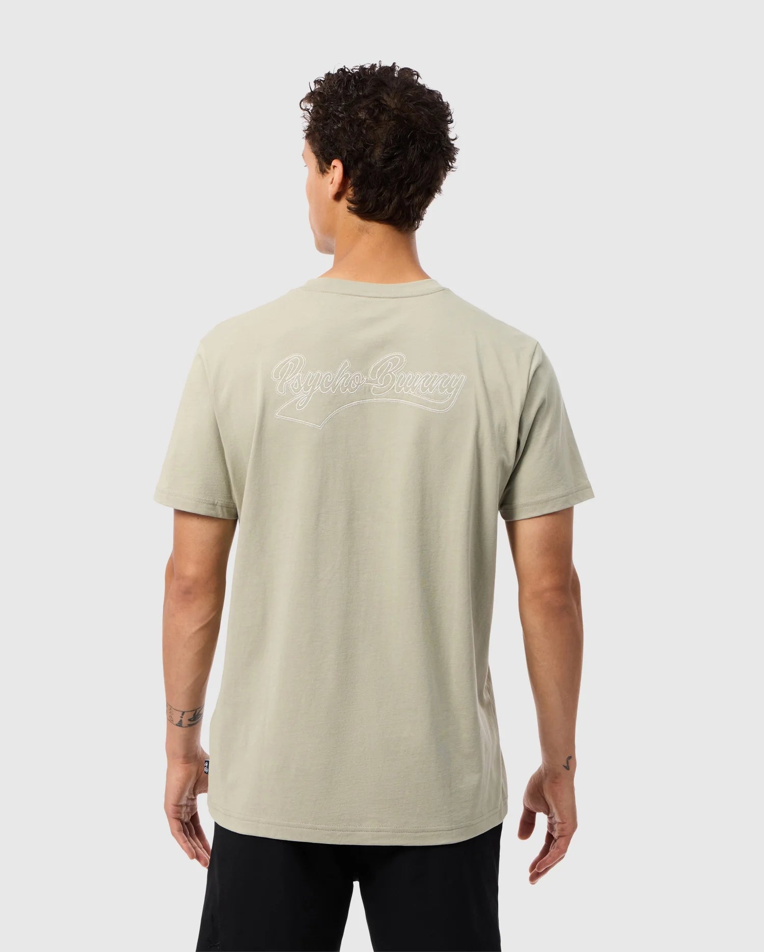A person with curly dark hair is wearing a light grey MENS PEARLAND BACK GRAPHIC TEE - B6U632C200 by Psycho Bunny, which has "Rogue Company" embroidered in white on the back. The person is depicted from behind with their left hand relaxed by their side, and they are dressed in black pants. The background is plain white.