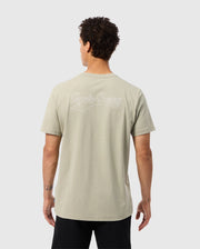 A person with curly dark hair is wearing a light grey MENS PEARLAND BACK GRAPHIC TEE - B6U632C200 by Psycho Bunny, which has "Rogue Company" embroidered in white on the back. The person is depicted from behind with their left hand relaxed by their side, and they are dressed in black pants. The background is plain white.