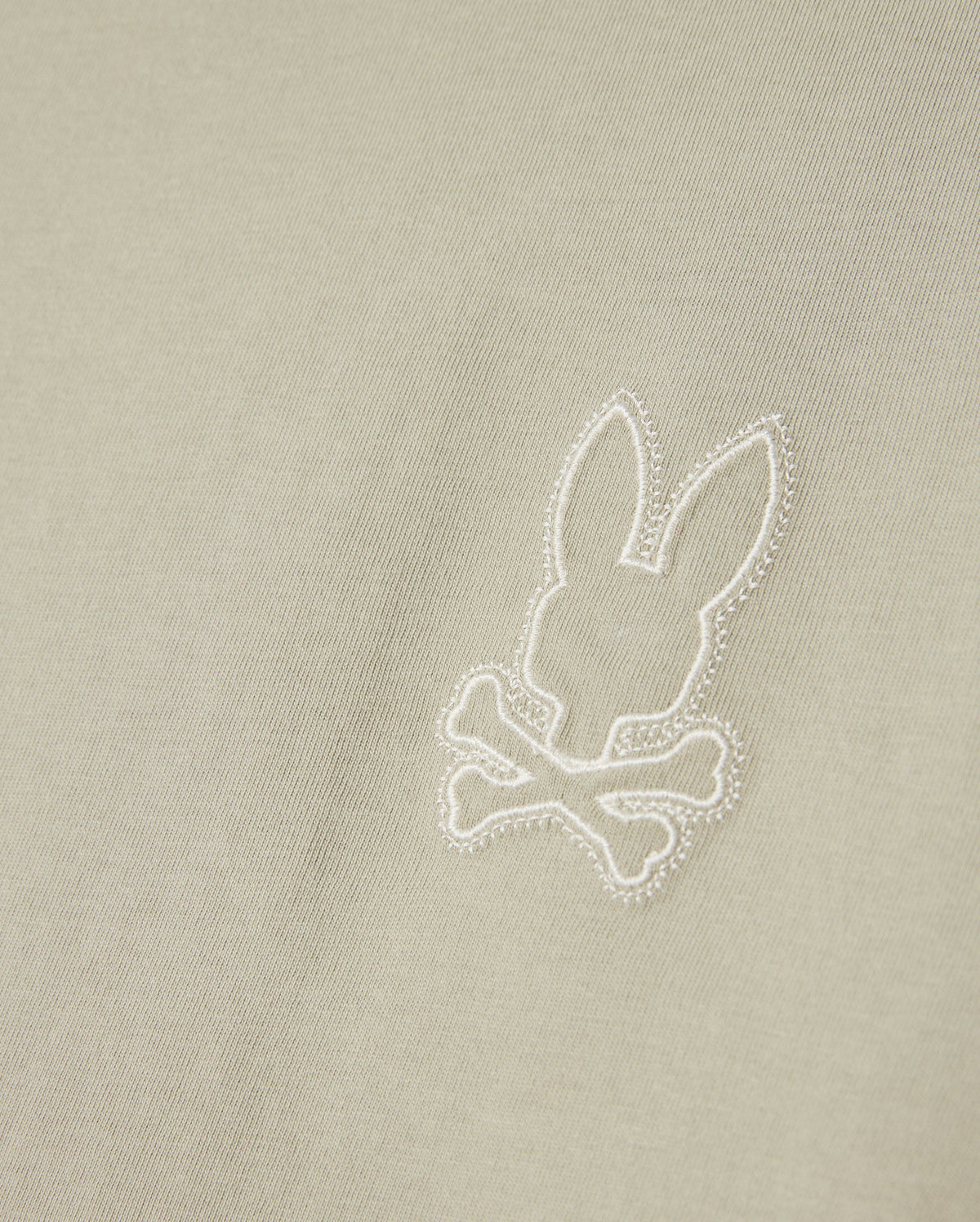 A close-up of light beige Pima cotton fabric from the Psycho Bunny MENS PEARLAND BACK GRAPHIC TEE - B6U632C200, featuring an embroidered design of a bunny head above crossed bones, reminiscent of a skull and crossbones motif. The embroidery is white, creating a subtle contrast against the high-quality Pearland tee.