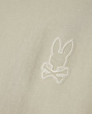 A close-up of light beige Pima cotton fabric from the Psycho Bunny MENS PEARLAND BACK GRAPHIC TEE - B6U632C200, featuring an embroidered design of a bunny head above crossed bones, reminiscent of a skull and crossbones motif. The embroidery is white, creating a subtle contrast against the high-quality Pearland tee.