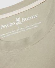 Close-up of the neckline of a beige MENS PEARLAND BACK GRAPHIC TEE - B6U632C200 showcasing the embroidered brand name "Psycho Bunny" beneath a bunny logo. The text includes "100% Pima Cotton," size "5," and "Made in Peru" written in English, French, and Spanish. The fabric features a ribbed texture.