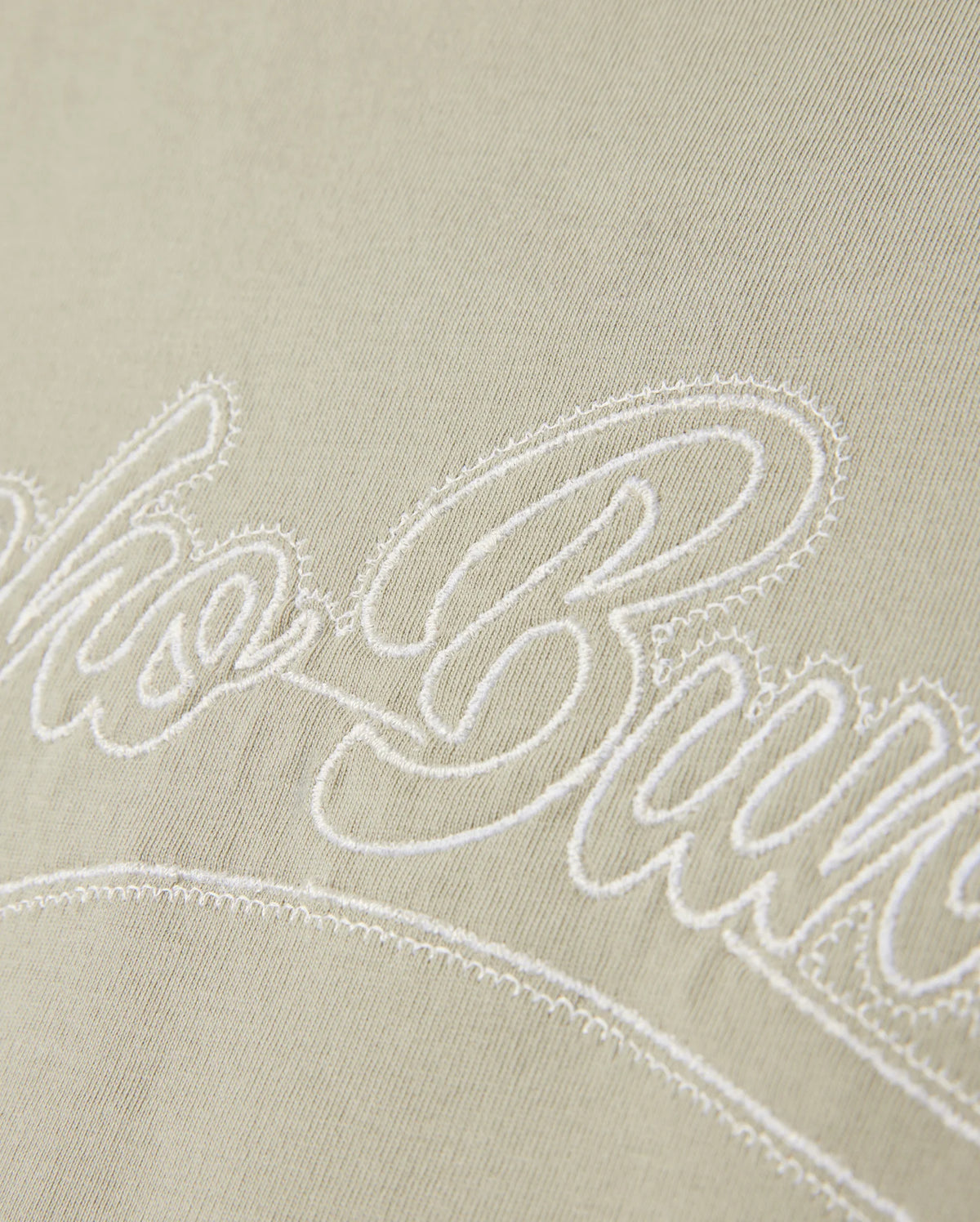 A close-up of the light Pima cotton fabric on the MENS PEARLAND BACK GRAPHIC TEE - B6U632C200 by Psycho Bunny, showcasing the white embroidered text "Club Bure" in a cursive font. The embroidery is slightly raised above the fabric surface, creating a textured effect.