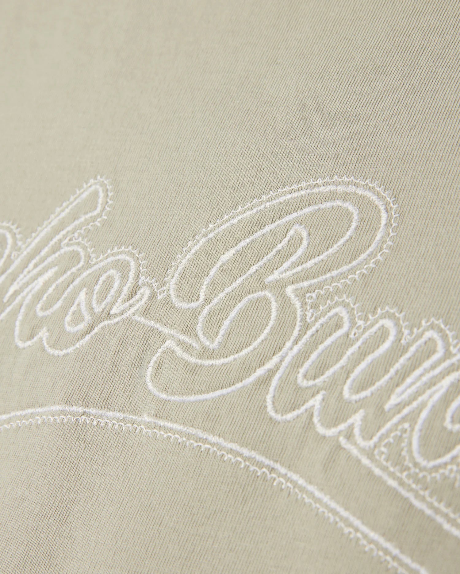 A close-up of the light Pima cotton fabric on the MENS PEARLAND BACK GRAPHIC TEE - B6U632C200 by Psycho Bunny, showcasing the white embroidered text "Club Bure" in a cursive font. The embroidery is slightly raised above the fabric surface, creating a textured effect.