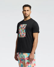 A man wears the Psycho Bunny MENS LEMMON GRAPHIC TEE - B6U655F200, a black Pima cotton T-shirt with a colorful abstract design, paired with multicolored patterned shorts for all-day comfort against a plain white backdrop.