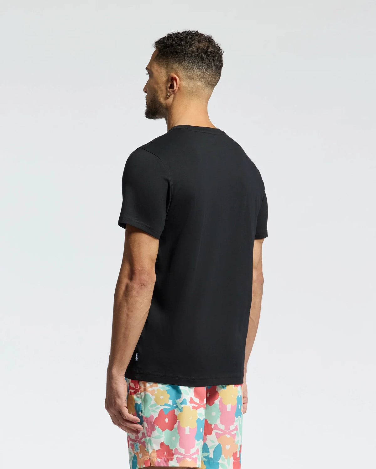 With short curly hair, the person stands facing away, wearing Psycho Bunny's MENS LEMMON GRAPHIC TEE - B6U655F200 and colorful floral shorts. The soft Pima cotton tee offers all-day comfort, complementing the scene's gentle gradient from light gray to white.