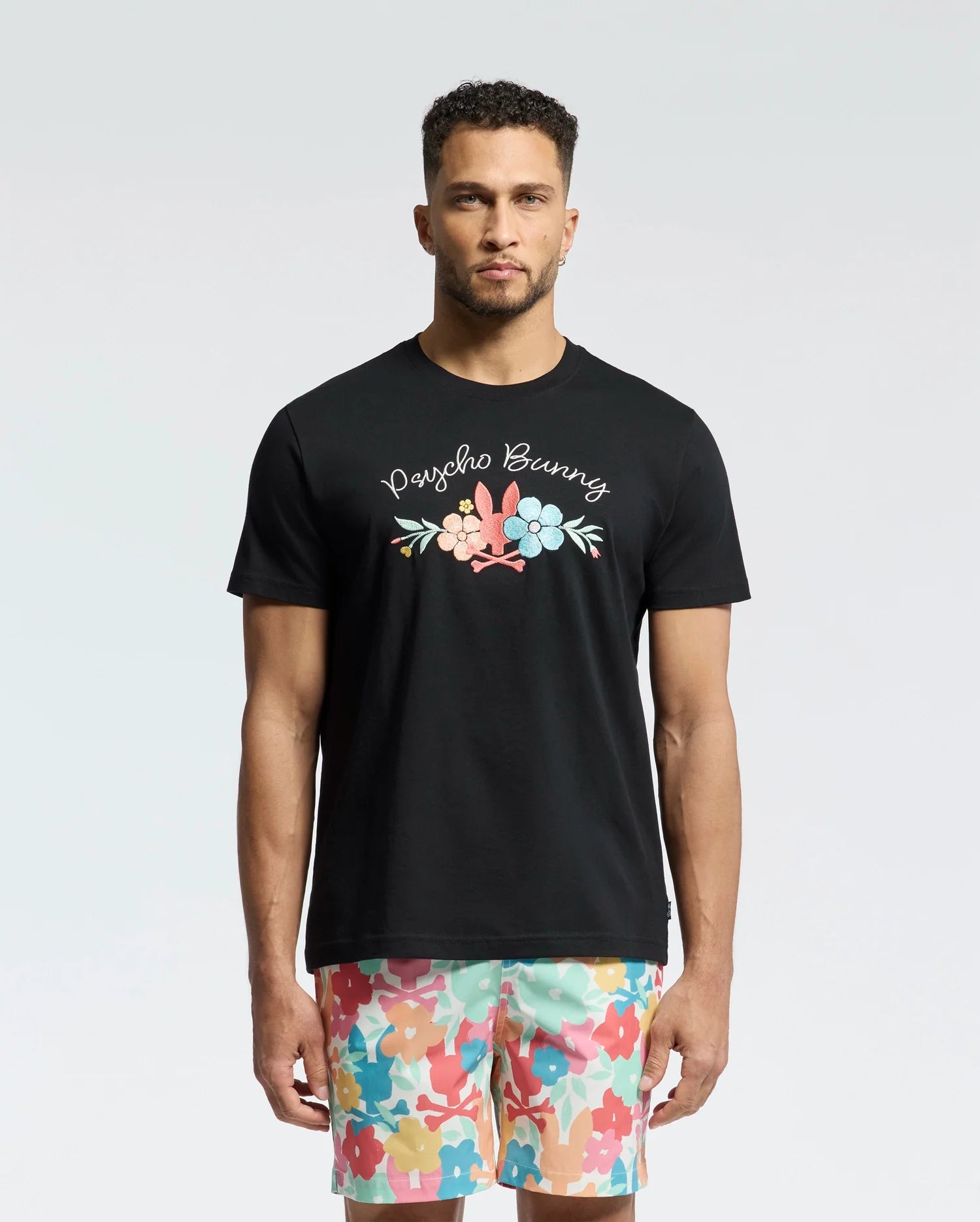 A man in a black Psycho Bunny MENS MEYLON GRAPHIC TEE - B6U656F200 with vibrant floral and text designs stands against a plain light background. His multicolored floral-patterned shorts enhance the casual flair of this Pima cotton ensemble, perfect for a sunny day out.