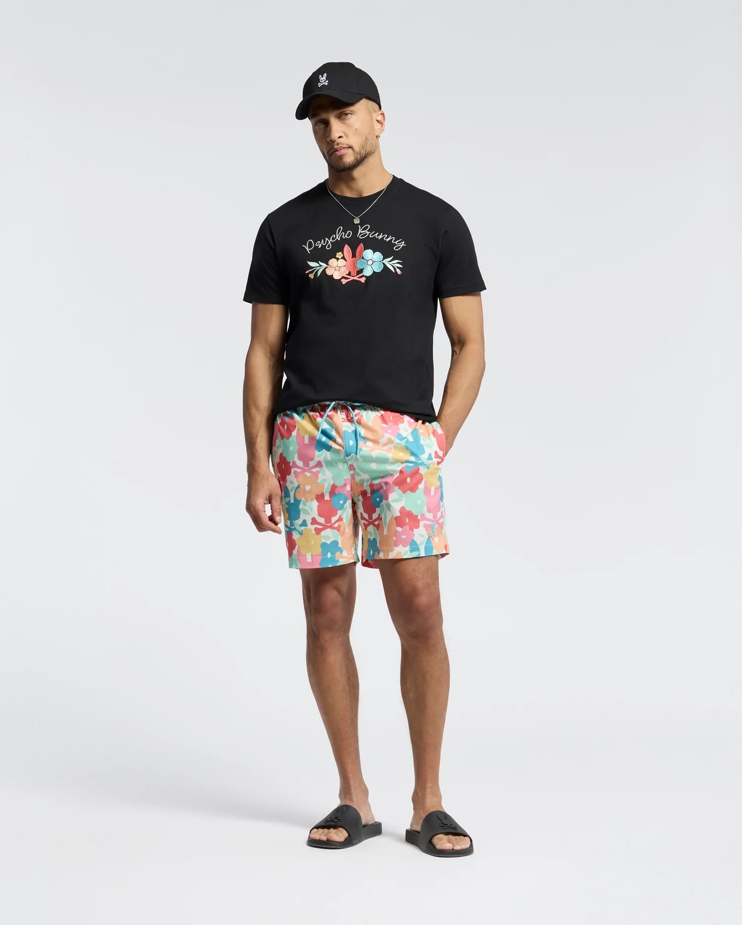 A man stands against a plain background wearing a black baseball cap, a Psycho Bunny MENS MEYLON GRAPHIC TEE - B6U656F200 with floral graphics, colorful floral-patterned shorts, and black slides.