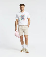 A man in a white Mens Lemora Graphic Tee by Psycho Bunny with a colorful print logo, beige shorts, white socks, beige sneakers, and holding a pink cap stands against a light gray background.