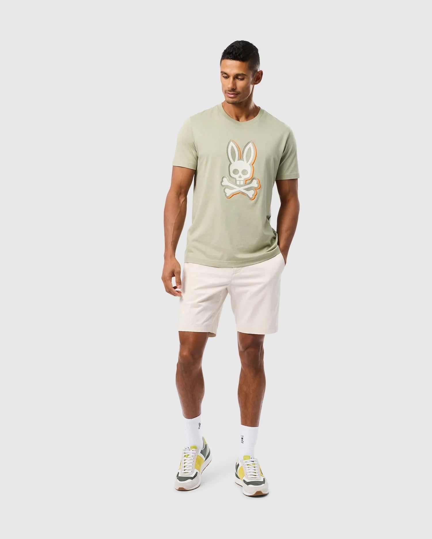 A man with short dark hair is wearing a light green **MENS KAYDEN GRAPHIC TEE - B6U676C200** featuring an embroidered bunny and crossbones motif by **Psycho Bunny**, paired with light beige shorts. He also has on white socks and green, white, and yellow sneakers. He is standing against a plain white background, looking downward.