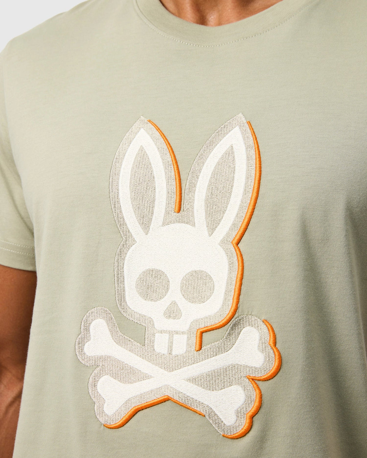 A person is wearing a light-colored MENS KAYDEN GRAPHIC TEE - B6U676C200 by Psycho Bunny, featuring an embroidered bunny's skull with crossbones beneath it. The design is outlined in orange, giving it a stylized appearance.