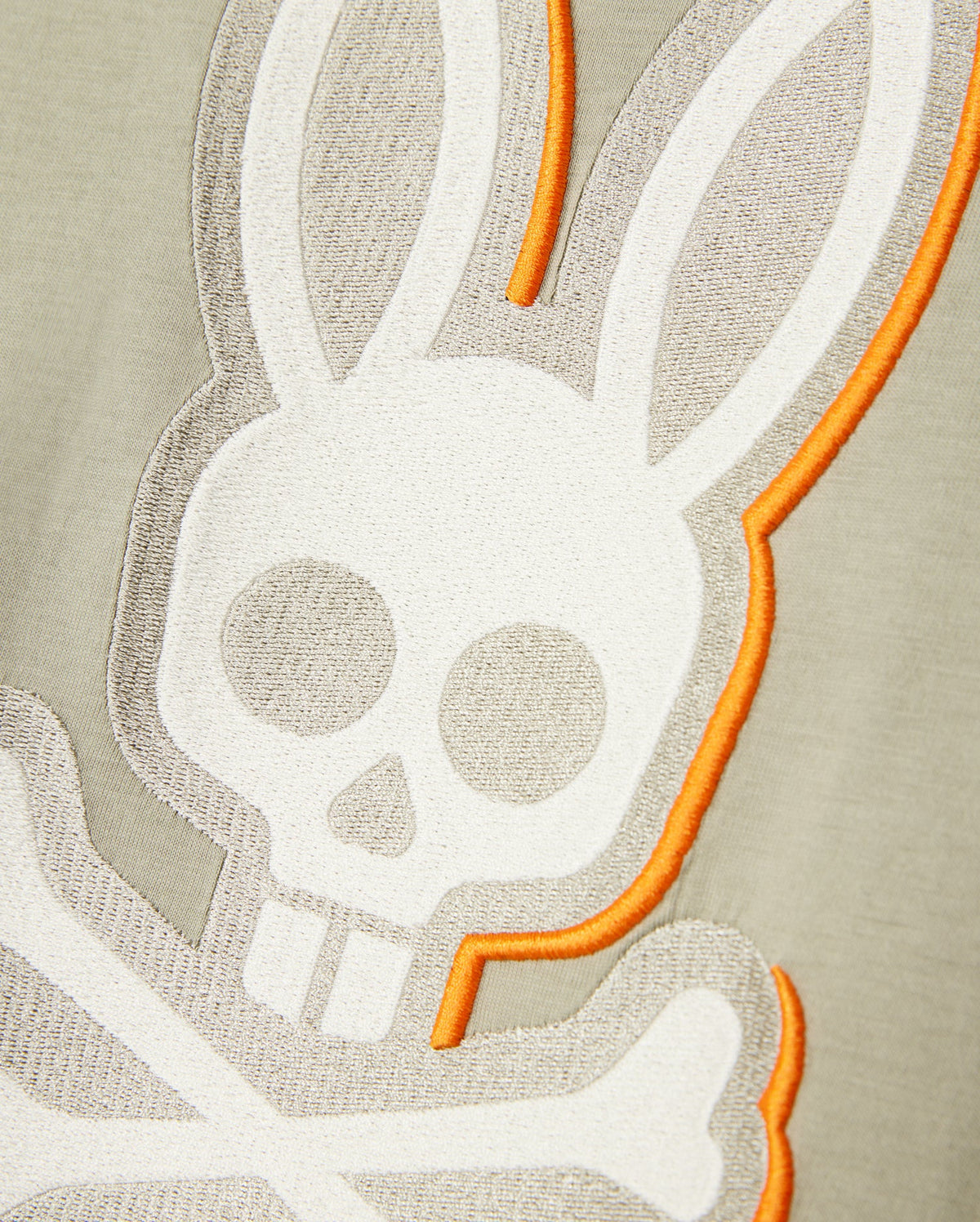 Close-up of an embroidered bunny design featuring a white skull and crossbones with bunny ears on a Psycho Bunny MENS KAYDEN GRAPHIC TEE - B6U676C200. The edges of the design are accentuated with orange stitching, enhancing the contrast against the Pima cotton's light gray and beige background.