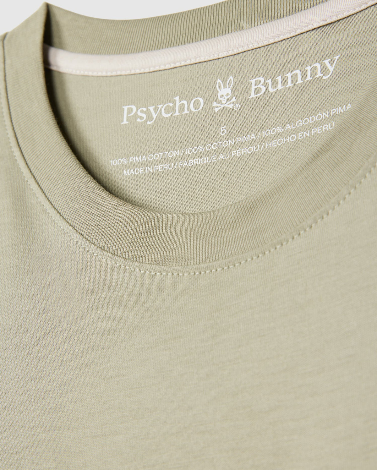 Close-up image of a light green MENS KAYDEN GRAPHIC TEE - B6U676C200. The label inside the neckline reads "Psycho Bunny," followed by an embroidered bunny logo and details about the material, indicating it is 100% Pima cotton and made in Peru.